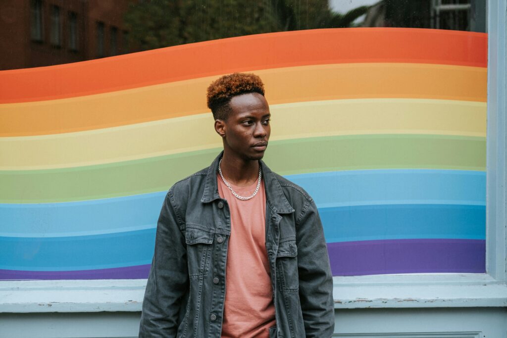 A thoughtful young man stands beside a rainbow mural, symbolizing diversity and inclusion.